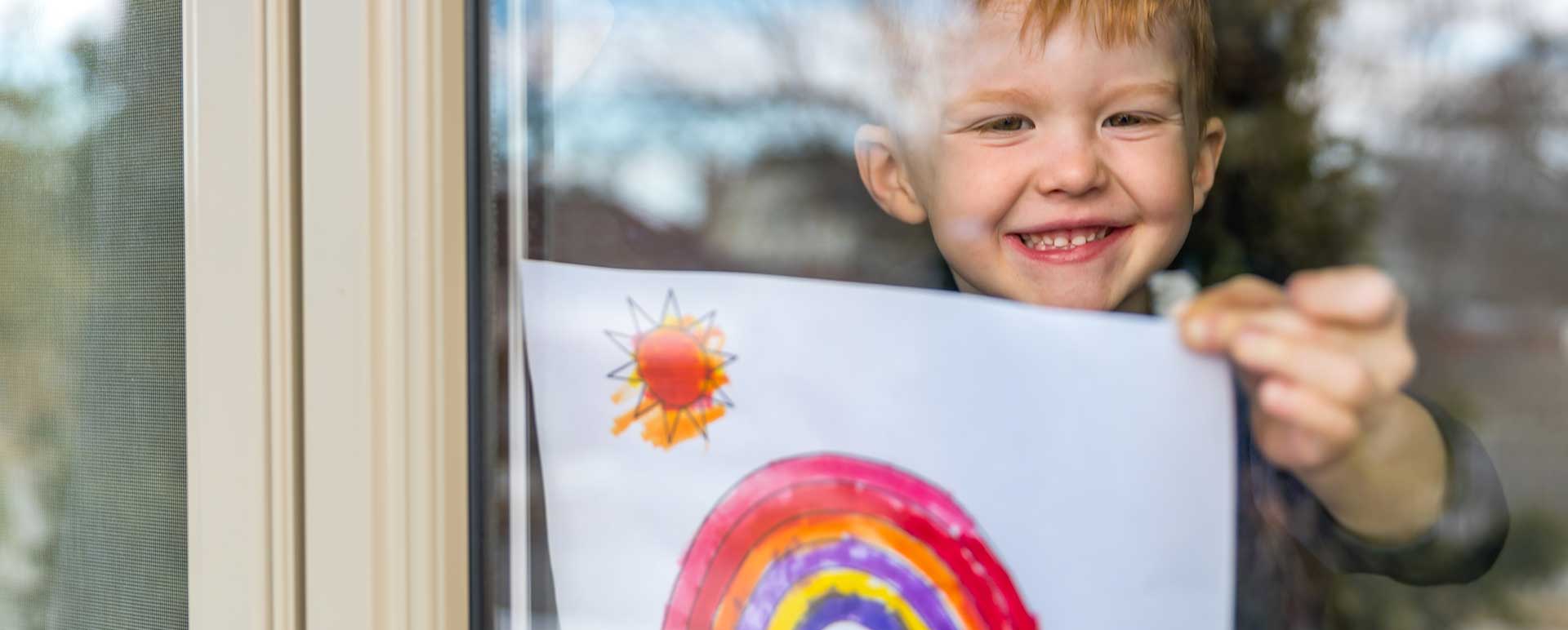 Boy holding up a drawing of a rainbow through a window
